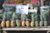 A market stall displays neatly arranged pineapples with a sign labeled 'Super Sweet MG-3'. Behind the pineapples, various other fruits such as apples are visible on a shelf. Nearby, there are metal containers, and a person is partly visible on the side.