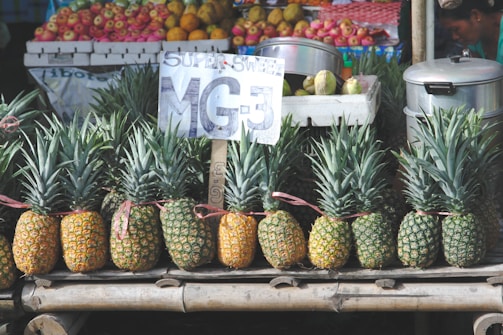 A market stall displays neatly arranged pineapples with a sign labeled 'Super Sweet MG-3'. Behind the pineapples, various other fruits such as apples are visible on a shelf. Nearby, there are metal containers, and a person is partly visible on the side.