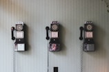 Three vintage rotary payphones are mounted on a light green textured wall, each with a black receiver hanging on its side.
