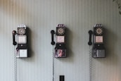 Three vintage rotary payphones are mounted on a light green textured wall, each with a black receiver hanging on its side.