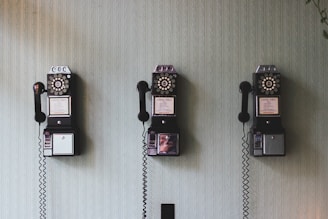 Three vintage rotary payphones are mounted on a light green textured wall, each with a black receiver hanging on its side.