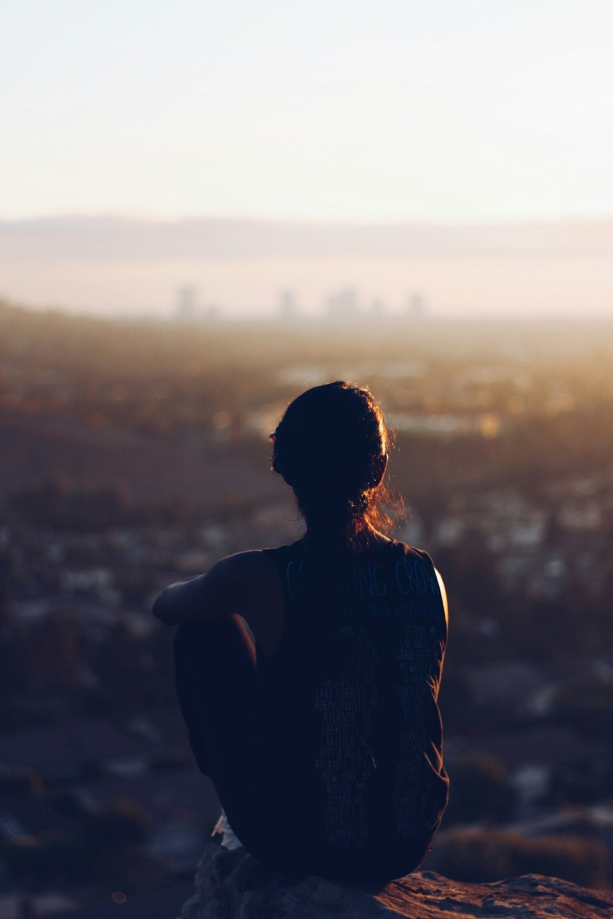 Woman seated on a brown rocky outcrop gazing toward mountains, contemplative natural-silence register